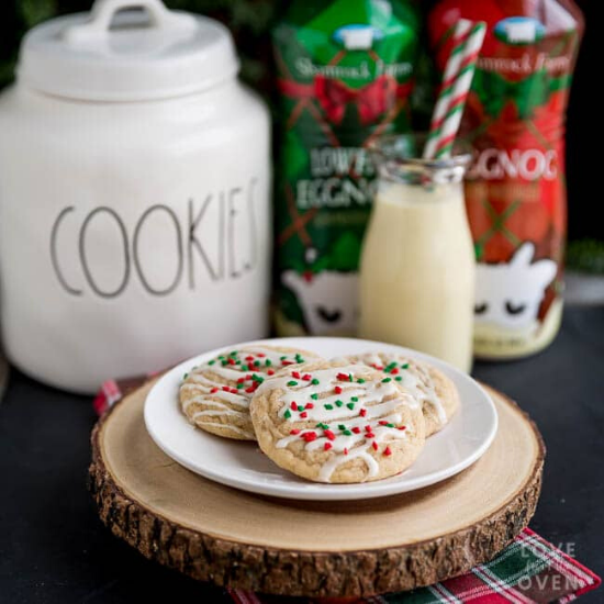 A plate of cookies styled on a wooden round with a cookies jar in the background alongside bottles of eggnog. 