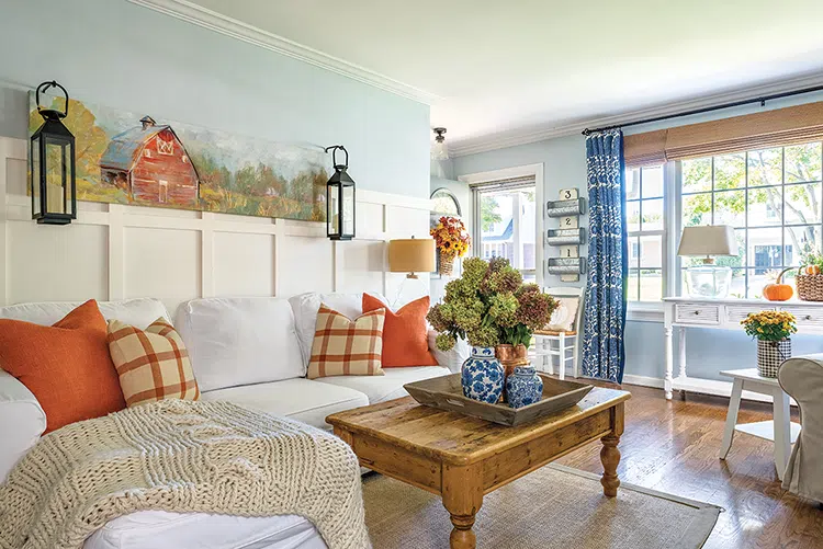 orange throw pillows and decorative pumpkin and mums, hydrangeas and sunflowers give fall feel to otherwise blue and white cozy cottage