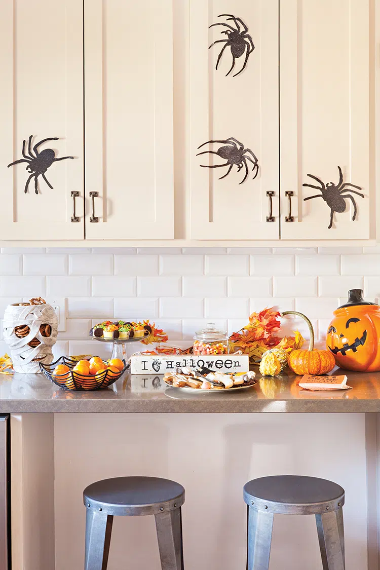 stick on spider stickers on cabinet fronts, mummy head shaped candy holder and spider's web style fruit bowl in home decorated for Halloween