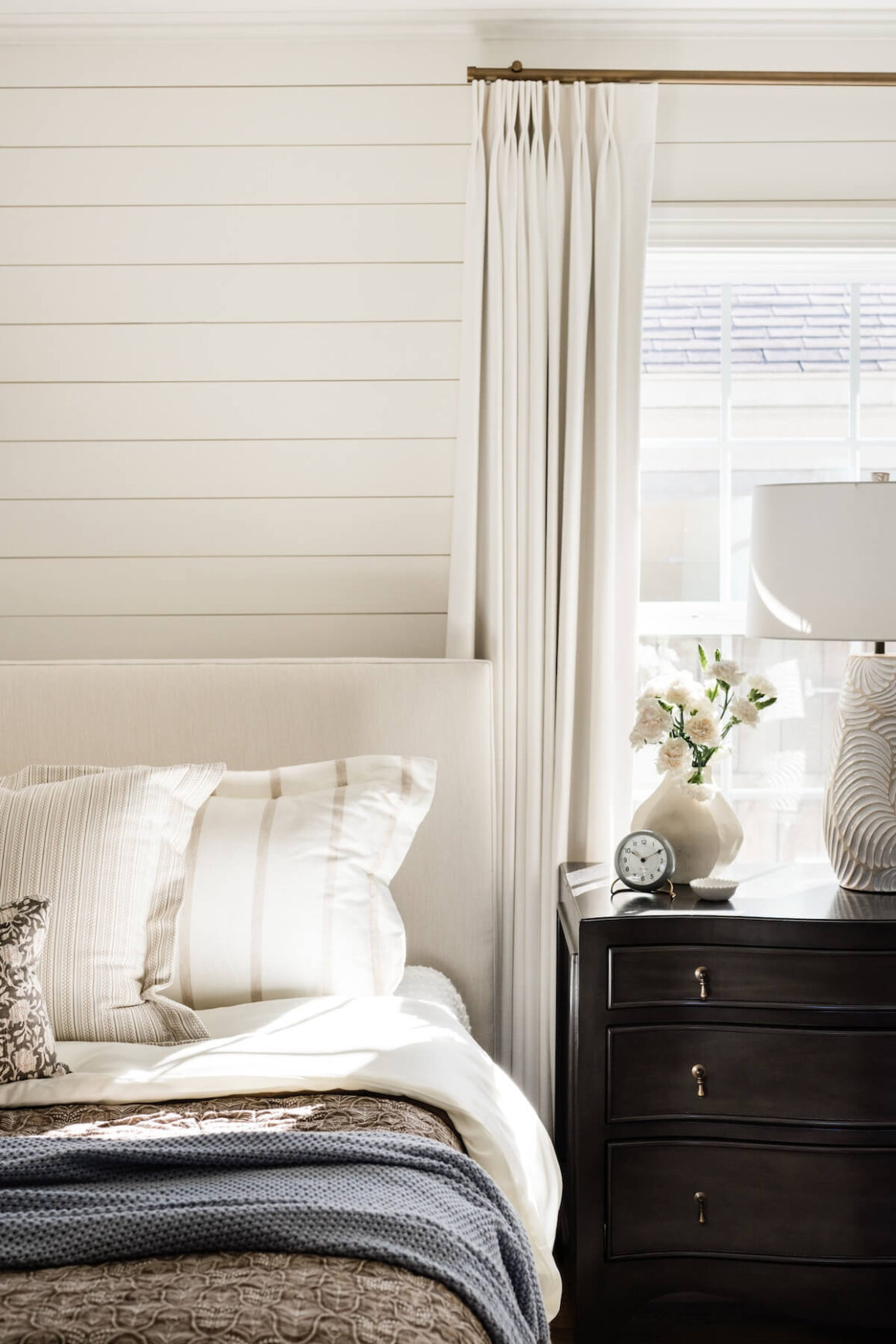 close up of bed with neutral and blue bedding textured lamp and dark wood nightstand