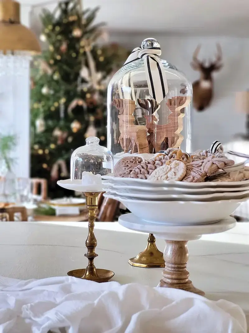 cloche covering wooden reindeer figurines and cake pedestal with bowl of cookie ornaments in neutral holiday home with natural accents