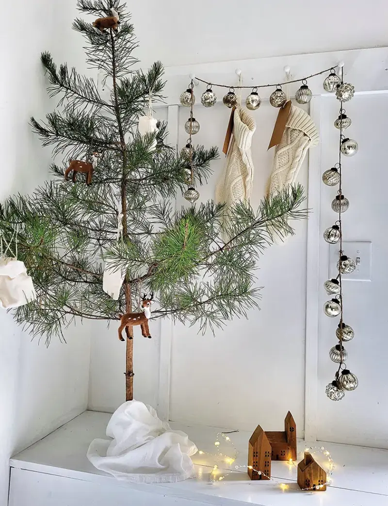 small Charlie Brown style tree with few ornaments next to garland of silver ornaments and white stockings on entryway bench and coat rack